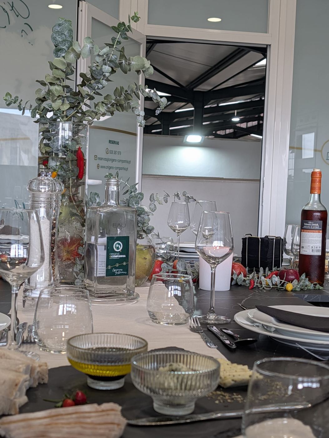 Mesa decorada com eucalipto, malaguetas e maçãs, copos, azeite e Moscatel de Setúbal, com a porta para o mercado ao fundo