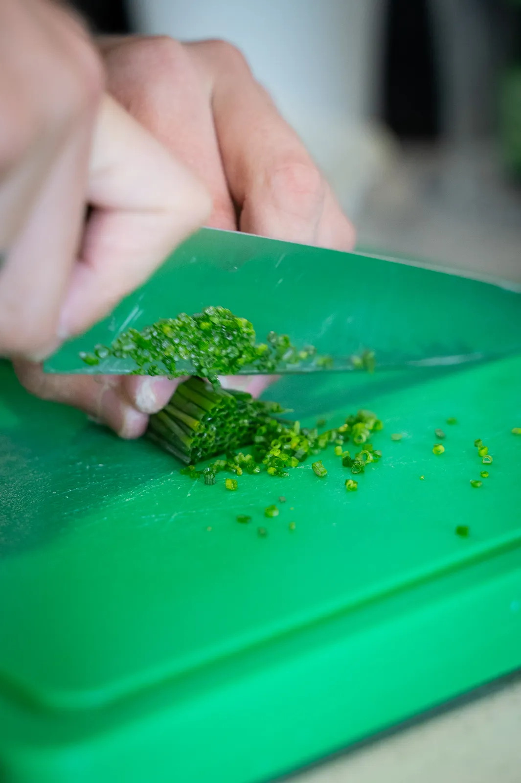 Hands finely chopping chives with a chef knife on a green cutting board